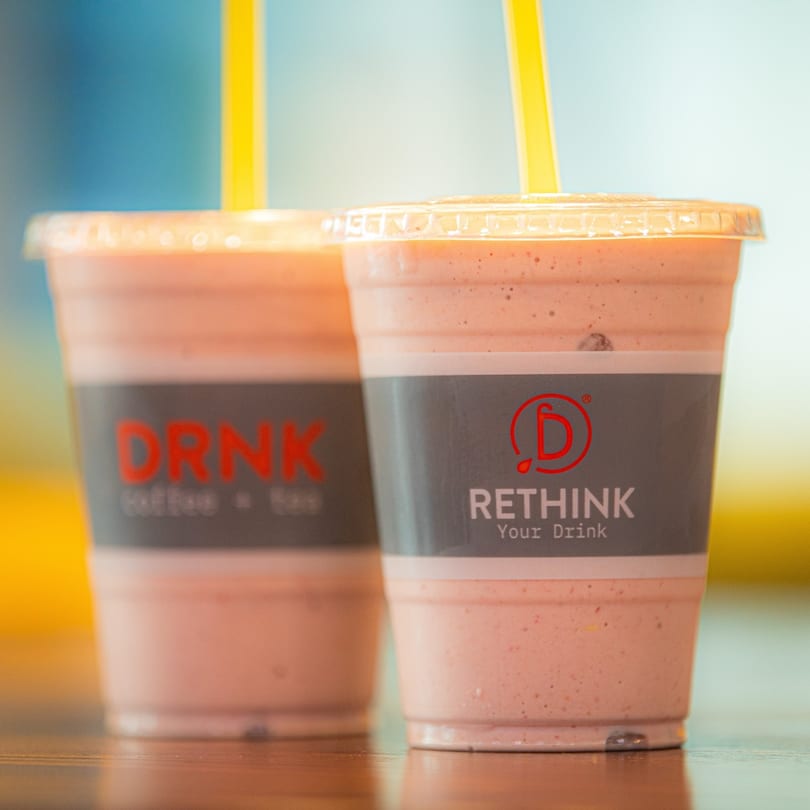 Two Qwench Juice Bar smoothies with yellow straws on a wooden surface.