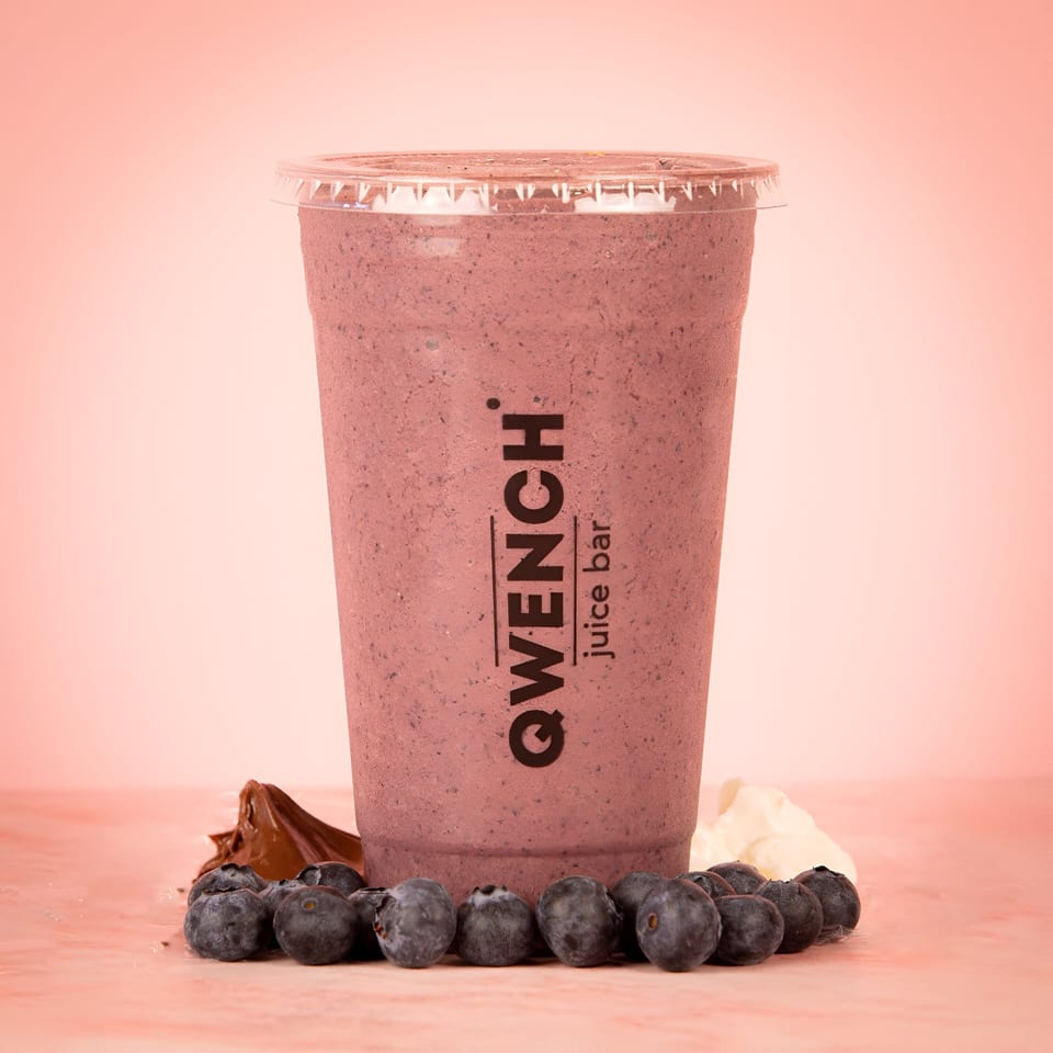 Qwench Juice Bar blueberry smoothie in a clear cup, surrounded by fresh blueberries and chocolate.