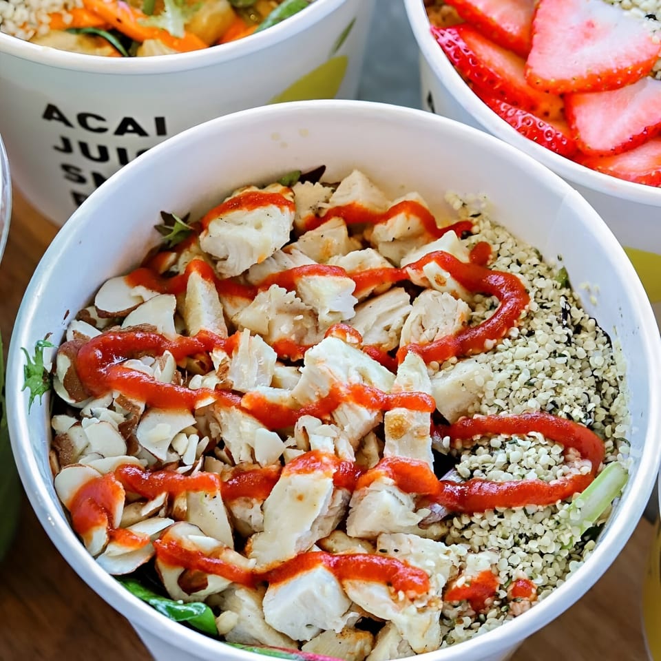 Close-up of a Qwench Juice Bar bowl with chicken, seeds, and red sauce, next to a strawberry bowl.