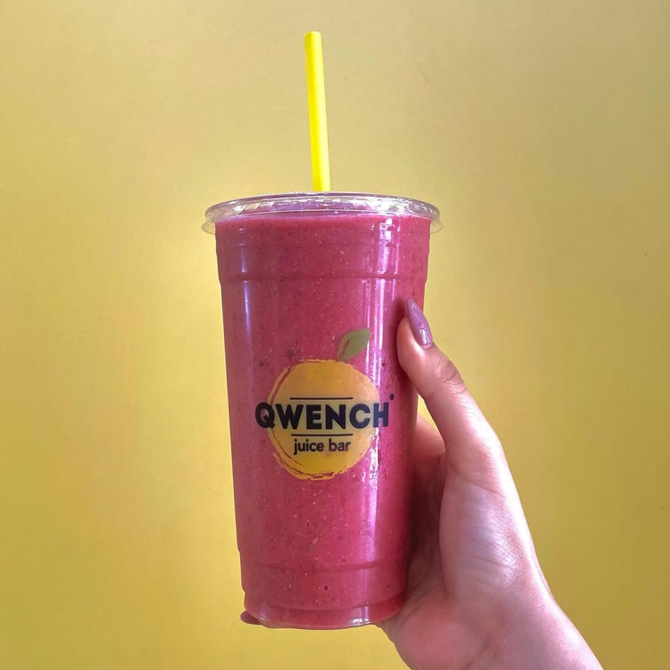 Hand holding a Qwench Juice Bar smoothie with a yellow straw against a yellow background.