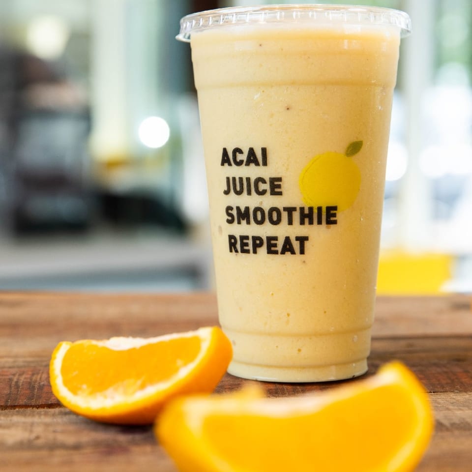 A Qwench Juice Bar smoothie with "ACAI JUICE SMOOTHIE REPEAT" text, next to orange slices on a wooden table.