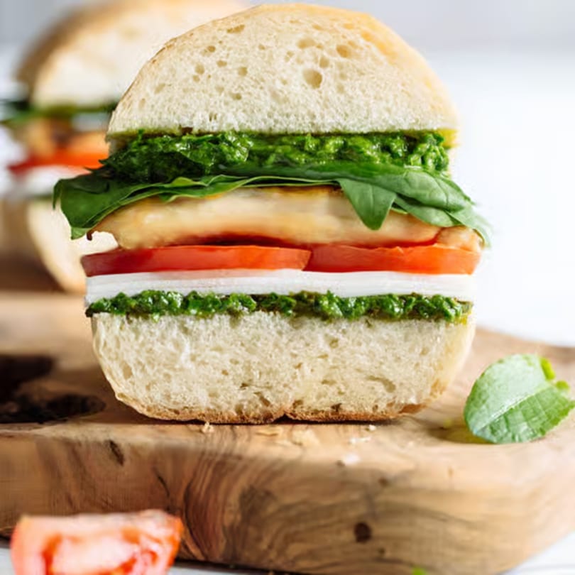 A close-up of a sandwich with pesto, mozzarella, tomato, and chicken on a wooden board.