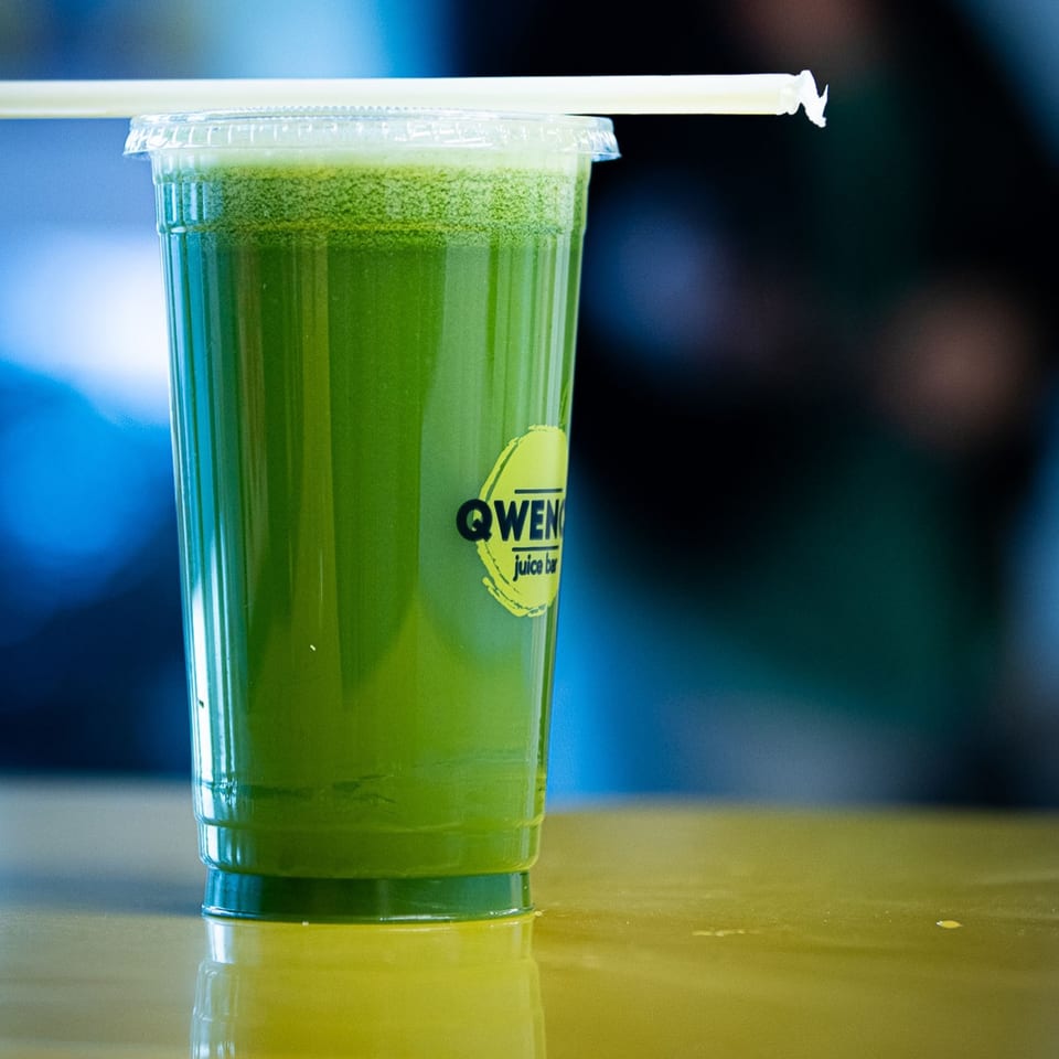 A Qwench Juice Bar green juice with a straw sits on a reflective surface.