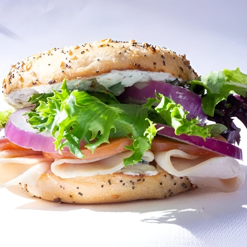 A bagel sandwich with turkey, lettuce, red onion, and cream cheese on a white background.